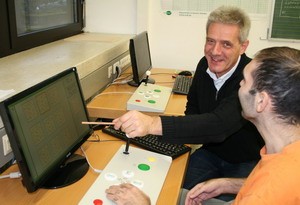 Zwei Personen sitzen an einem Tisch vor Computern. Eine Person, ein Mann mit grauen Haaren, zeigt mit einem Stift auf den Bildschirm und lächelt, während die andere Person aufmerksam zuschaut. Beide scheinen an einer gemeinsamen Aktivität oder Anleitung interessiert zu sein.