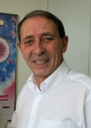 Ein lächelnder Mann in einem weißen Hemd steht vor einer farbigen Wand mit abstrakten Mustern. Sein Gesichtsausdruck ist freundlich und einladend.