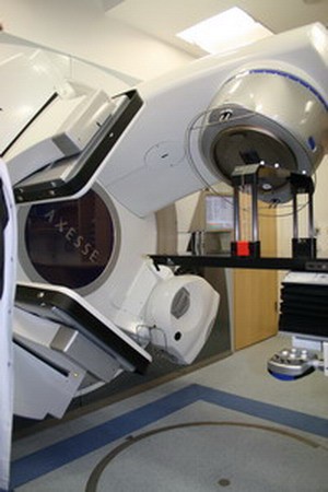 Bildbeschreibung: Ein modernes medizinisches Gerät zur Strahlentherapie in einem Behandlungsraum. Das Gerät hat eine große, bewegliche Struktur mit einem runden Element, das für die präzise Bestrahlung von Patienten ausgelegt ist. Im Hintergrund sind eine Wand und einige Möbel zu sehen.