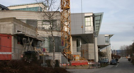 Eine Baustelle ist zu sehen, mit einem Kran, der funktioniert. Links steht ein unfertiges Gebäude, während rechts ein modernes, verglastes Gebäude erkennbar ist. Die Straße vorne ist teilweise befahrbar. Einige Bauprodukte und Absperrungen sind sichtbar.