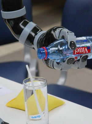 Ein roboterarm mit einer greifenden Hand hält eine Wasserflasche über ein Glas mit Wasser. Der Arm ist mechanisch und zeigt technische Details, während das Glas und die Flasche im Fokus stehen.
