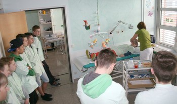 Eine Gruppe von Jugendlichen beobachtet einen medizinischen Eingriff oder eine Behandlung in einem Krankenhauszimmer. Eine Fachkraft kümmert sich um einen Patienten, der auf einem Krankenbett liegt. Die Umgebung ist sauber und praktisch gestaltet.