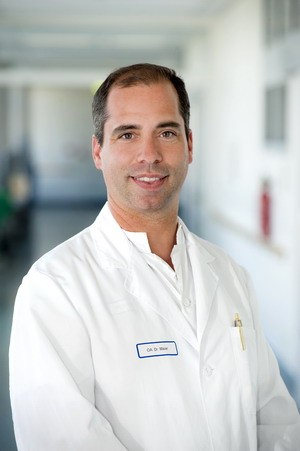 Ein männlicher Arzt in einem weißen Kittel lächelt freundlich in die Kamera. Im Hintergrund sind unscharfe Krankenhausflure zu sehen. Der Arzt trägt ein Namensschild und wirkt kompetent und einladend.