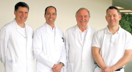 Vier Männer in weißen Laborkitteln stehen lächelnd nebeneinander. Sie scheinen medizinisches Fachpersonal zu sein. Der Hintergrund ist hell und neutral, was den Fokus auf die Personen lenkt.