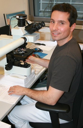 Ein Mann sitzt an einem Tisch, der mit einem Mikroskop und wissenschaftlichen Unterlagen ausgestattet ist. Er lächelt freundlich in die Kamera und trägt ein dunkelgraues T-Shirt. Im Hintergrund sind Fenster zu sehen, durch die Tageslicht fällt.