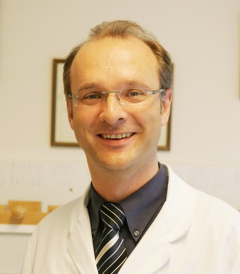 Ein lächelnder Arzt in weißem Kittel und mit Brille steht vor einer neutralen Hintergrundwand. Er trägt ein dunkles Hemd und eine gestreifte Krawatte. Der Gesichtsausdruck ist freundlich und einladend.