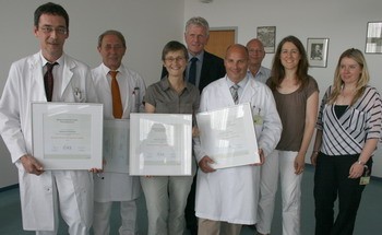 Gruppenschärfe in einem Krankenhaus. Acht Personen in weißer Berufskleidung halten Urkunden. Sie zeigen Stolz und Freude über ihre Auszeichnungen. Der Hintergrund zeigt eine Büroumgebung mit wenigen weiteren Personen.