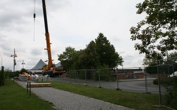 Ein großer Baukran steht auf einer Baustelle, umgeben von einem Bauzaun und Bäumen. Im Hintergrund ist ein Gebäude mit einer markanten Dachform sichtbar. Der Weg ist mit Kieselsteinen bedeckt, der in Richtung des Krans führt.