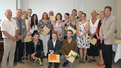 Eine Gruppe von etwa 20 Personen steht im Raum und lächelt. Einige halten Urkunden und Blumen in den Händen. Sie scheinen anlässlich einer Feier oder Abschlusszeremonie versammelt zu sein. Die Atmosphäre wirkt festlich und fröhlich.