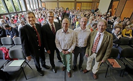 Eine Gruppe von fünf Männern steht im Vordergrund, während im Hintergrund eine große Menschenmenge in einem Raum sitzt. Die Atmosphäre wirkt informell und lebhaft, was auf eine Veranstaltung oder eine Ansprache hindeutet.