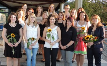 Eine Gruppe von 16 Frauen steht lächelnd auf einer Treppe. Einige halten Sonnenblumen und Blumensträuße in den Händen. Sie sind in verschiedenen Kleidungsstilen gekleidet, darunter festliche und legere Outfits. Die Umgebung wirkt freundlich und feierlich, möglicherweise zu einer besonderen Anlässe.