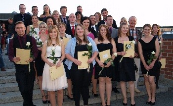 Eine Gruppe von Absolventen steht auf einer Treppe, lächelt und hält Urkunden in den Händen. Einige Teilnehmer tragen formelle Kleidung, während andere Kleider oder Anzüge mit weißen Rosen in den Händen tragen. Der Hintergrund zeigt eine festliche Atmosphäre.