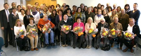 Gruppenfoto mit etwa 50 Personen, die überwiegend sitzen. Viele der Anwesenden halten bunte Blumensträuße in den Händen. Einige stehen im Hintergrund. Die Atmosphäre wirkt festlich und freundlich, wahrscheinlich anlässlich eines besonderen Ereignisses oder einer Feier.