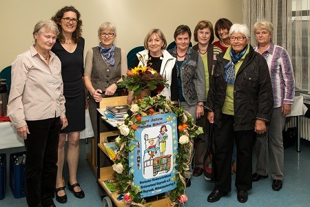 Eine Gruppe von zehn Frauen steht lächelnd vor einem Bücherwagen, der mit Blumen dekoriert ist. Auf dem Wagen befindet sich ein Plakat, das 30 Jahre einer Initiative feiert. Die Frauen wirken stolz und zeigen eine freundliche Atmosphäre.
