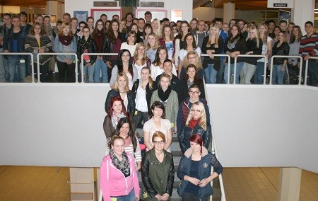 Eine große Gruppe von Schülern steht auf einer Treppe in einem Gebäude. Die Personen sind in mehreren Reihen angeordnet und lächeln. Die Gruppe besteht aus überwiegend jungen Erwachsenen, die diverse Kleidung tragen.