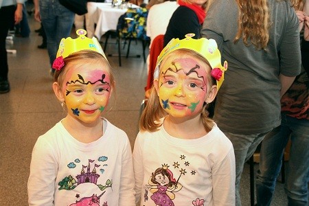 Zwei Mädchen mit fröhlichen, bemalten Gesichtern stehen nebeneinander. Jedes trägt eine gelbe Krone mit Blumen. Die Mädchen tragen T-Shirts mit kindlichen Motiven. Die Umgebung zeigt verschwommen andere Menschen und Tische, was auf eine feierliche Veranstaltung hindeutet.