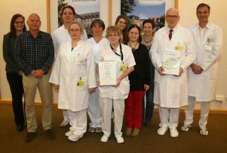Eine Gruppe von zehn Personen in Krankenhauskleidung steht in einem Raum. Einige halten Zertifikate in der Hand. Es gibt eine Mischung aus männlichen und weiblichen Teammitgliedern, die lächeln. Der Hintergrund zeigt Poster an der Wand.
