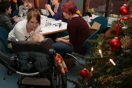 Eine Gruppe von Menschen sitzt an einem Tisch und bastelt. Im Vordergrund ist ein Kinderwagen mit einem kleinen Kind zu sehen. Im Hintergrund steht ein Weihnachtsbaum mit dekorierten Kugeln und Kerzen, was eine festliche Atmosphäre schafft.