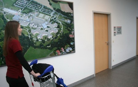 Eine Frau mit einem Gepäckwagen steht in einem Flur und schaut auf eine große Luftaufnahme einer Anlage an der Wand. Rechts führt eine Tür in einen anderen Raum. Der Flur ist hell und modern gestaltet.