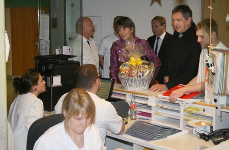 Eine Gruppe von Menschen steht an einem Empfangstresen in einer Einrichtung. Ein Mann überreicht einen Geschenkkorb, während mehrere Personen miteinander sprechen. Einige Mitarbeiter sind mit Computern beschäftigt. Die Atmosphäre wirkt freundlich und festlich.