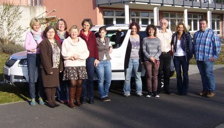 Eine Gruppe von zehn Personen steht lächelnd vor einem weißen Auto, vor einem Gebäude. Sie sind unterschiedlich gekleidet und scheinen sich gut zu verstehen. Die Szene vermittelt ein Gefühl von Gemeinschaft und Freude.