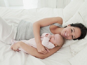 Eine glückliche Mutter liegt auf einem Bett und hält ihr schlafendes Baby in den Armen. Beide strahlen Zufriedenheit und Geborgenheit aus. Das Bild vermittelt eine liebevolle Verbindung zwischen Mutter und Kind.