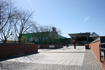 Das Bild zeigt einen gepflasterten Weg vor einem modernen Gebäude mit einem grünen Fassadenbereich. Menschen gehen entlang des Weges, bei klarem Himmel und Sonnenschein. Einige Bäume sind im Hintergrund sichtbar.