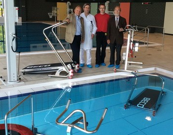 Vier Männer stehen am Beckenrand eines Schwimmbades. Im Hintergrund sind verschiedene Geräte und eine Rampe zu sehen, die wahrscheinlich für die Unterstützung von Menschen mit eingeschränkter Mobilität gedacht sind.