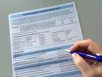Das Bild zeigt ein Patientenfragebogen der Klinik Bayreuth GmbH. Eine Hand hält einen Stift und markiert Kästchen auf dem Formular. Der Fragebogen enthält allgemeine Fragen zur Patientenerfahrung und zur Zufriedenheit mit den Dienstleistungen der Klinik.