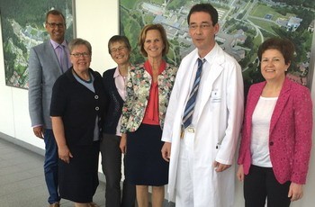 Sechs Personen stehen lächelnd in einer modernen Umgebung, einige in formeller Kleidung, darunter ein Arzt in einem weißen Kittel. Im Hintergrund sind große Wandbilder zu sehen. Die Gruppe scheint eine positive Atmosphäre zu vermitteln.