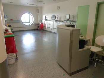 Eine helle, moderne Krankenhausumgebung mit einem Empfangstresen im Vordergrund. An der Wand befinden sich mehrere medizinische Geräte. Ein großes rundes Fenster lässt Tageslicht herein. Die Böden sind hell und der Raum wirkt aufgeräumt und sauber.