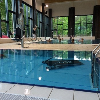 Ein Hallenbad mit einer großen Fensterfront, durch die viel Tageslicht strömt. Im Wasser sind mehrere Personen zu sehen, die sich im Wasser bewegen oder Übungen machen. Am Rand des Pools sind Fitnessgeräte platziert.