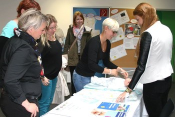 Eine Gruppe von Frauen schaut zu, während eine Frau an einem Tisch zeigt, wie man ein Baby wickelt. Auf dem Tisch liegen Babyutensilien und Informationsmaterialien. Die Atmosphäre ist lehrreich und aufmerksam.