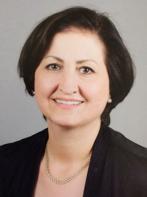 Das Bild zeigt eine lächelnde Frau mit kurzem, dunklem Haar. Sie trägt eine schwarze Jacke und eine kleine Perlenkette. Der Hintergrund ist grau. Die Frau sieht freundlich und sympathisch aus.