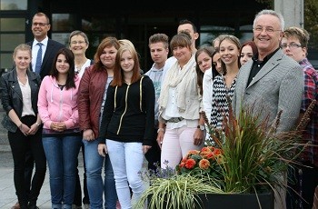 Eine Gruppe von Jugendlichen steht in einem Außenbereich, umgeben von Pflanzen. Einige von ihnen lächeln, während mehrere Erwachsene, darunter ein Mann in einem Anzug, ebenfalls dabei sind. Die Szenerie vermittelt eine freundliche und einladende Atmosphäre.