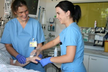 Zwei Krankenschwestern in blauen Kitteln und Handschuhen lächeln, während sie sich um einen Patienten kümmern. Eine Schwester führt eine Pflegehandlung an dem Arm des Patienten aus, während die andere sie freundlich beobachtet. Die Szene strahlt Professionalität und Fürsorge aus.