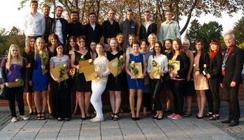 Eine Gruppe von circa 30 Personen steht im Freien zusammen, um einen feierlichen Moment zu teilen. Einige halten Zertifikate in der Hand, während sie Blumen tragen. Die Mehrheit ist in formeller Kleidung gekleidet. Im Hintergrund sind Bäume und eine Abendstimmung sichtbar.